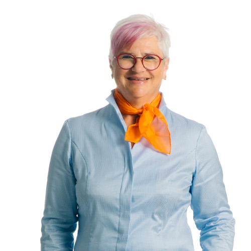 Eine ältere Frau mit rosa gefärbtem Haar, runden Brillen und einem orangefarbenen Schal lächelt in die Kamera.